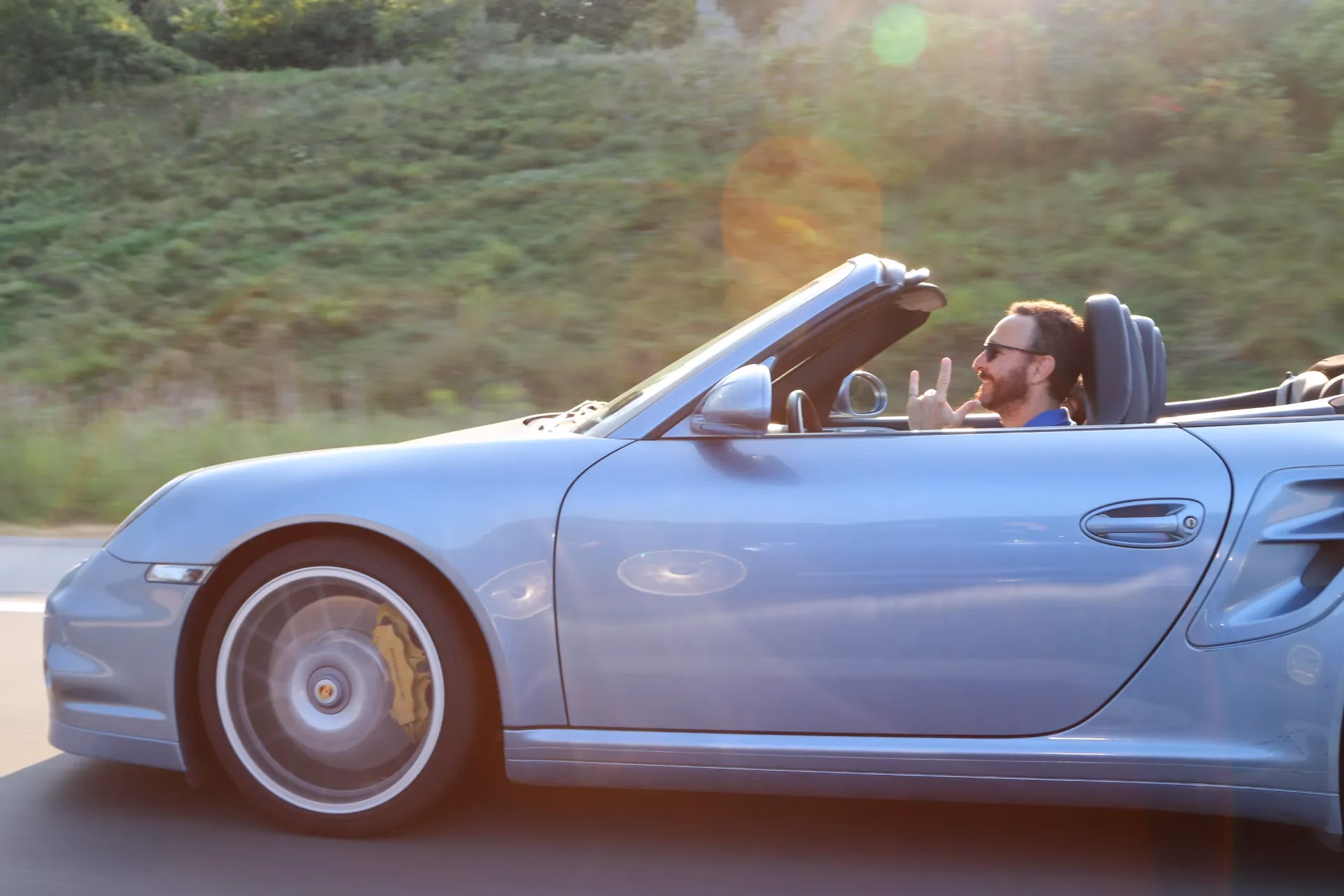 A light blue Porsche 911 cabriolet on the road, the driver waving to the camera