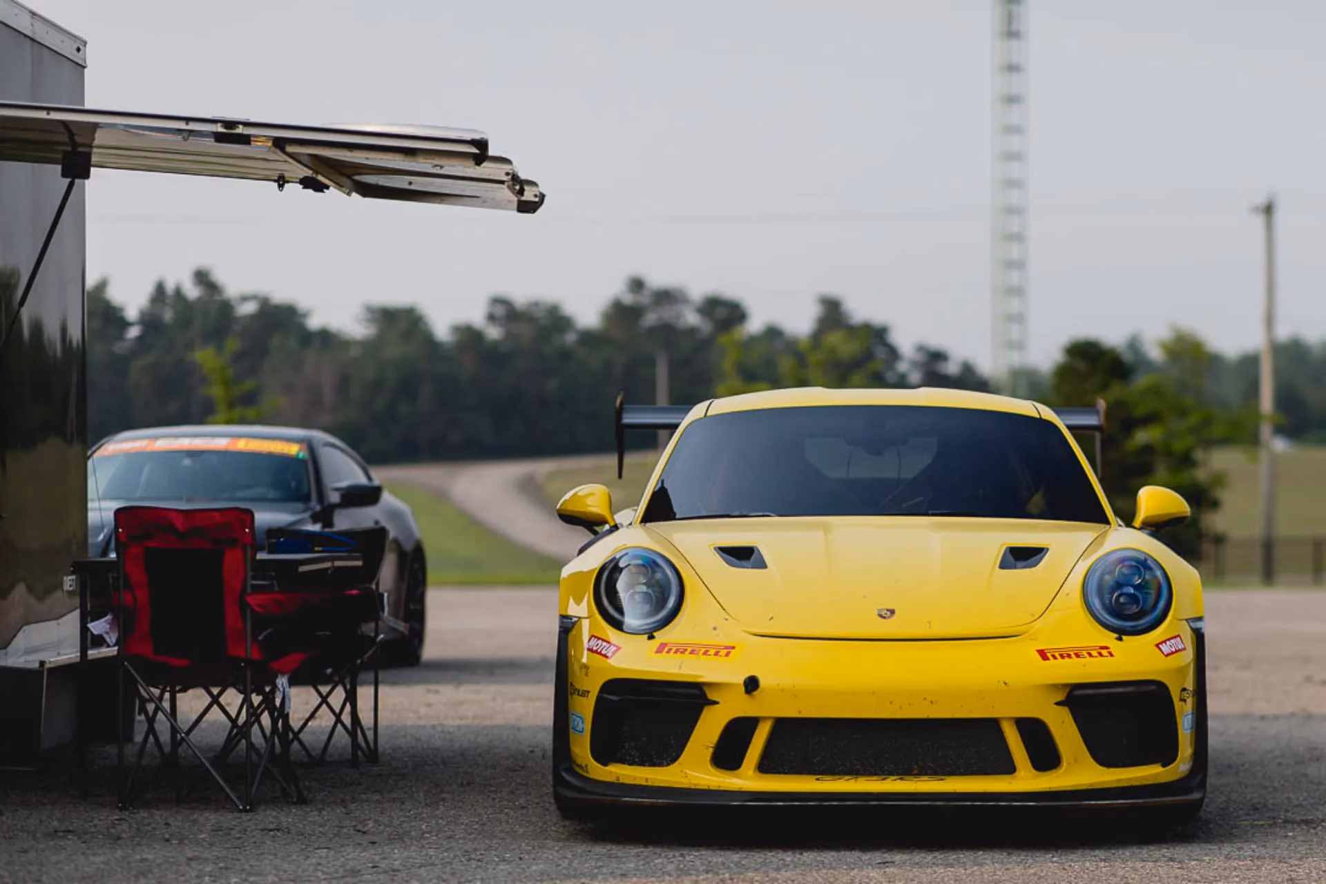 A yellow Porsche 911 GT3 parked in the paddock beside a trailer and camp chairs