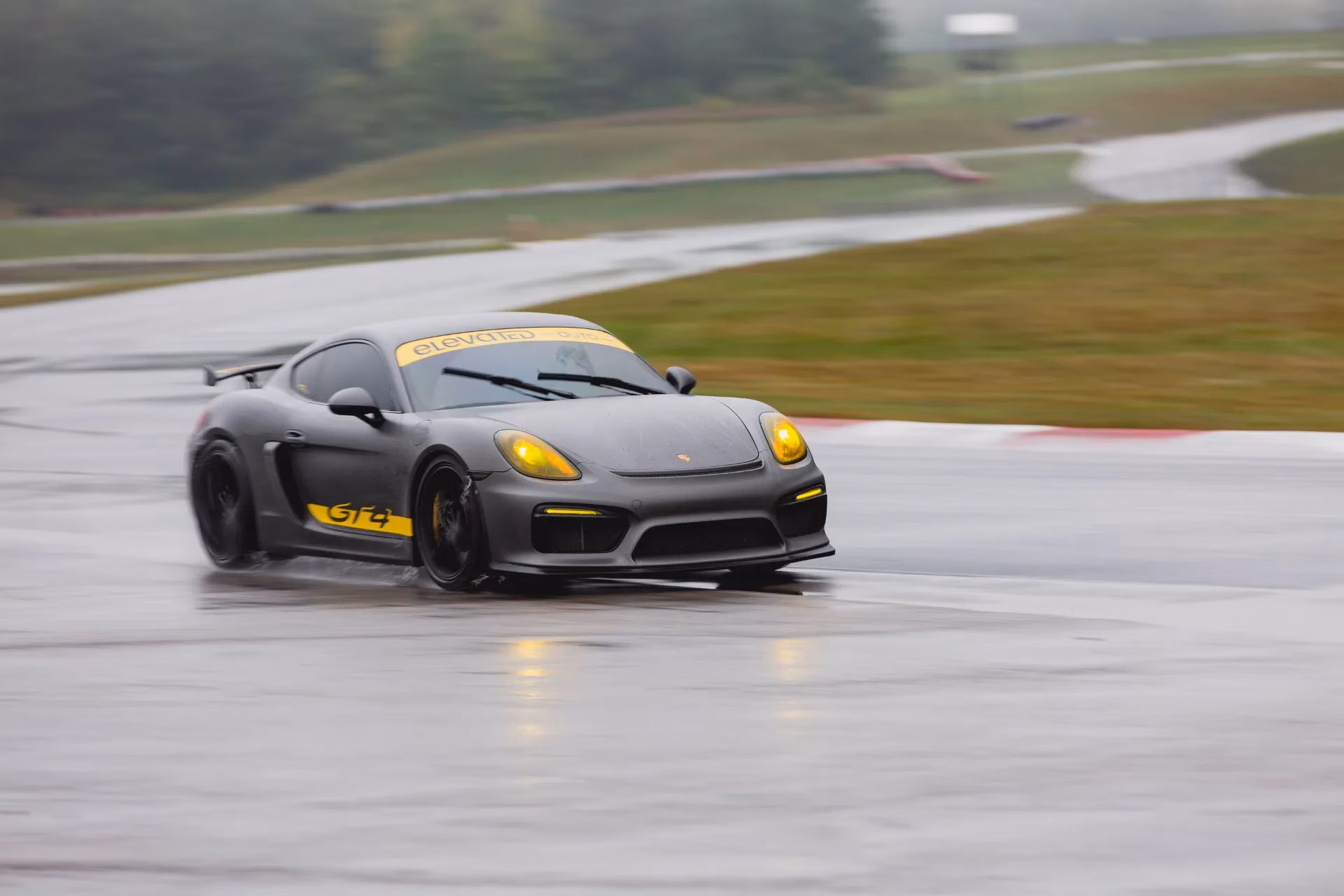 A grey Porsche Cayman GT4 with yellow accents on a wet track