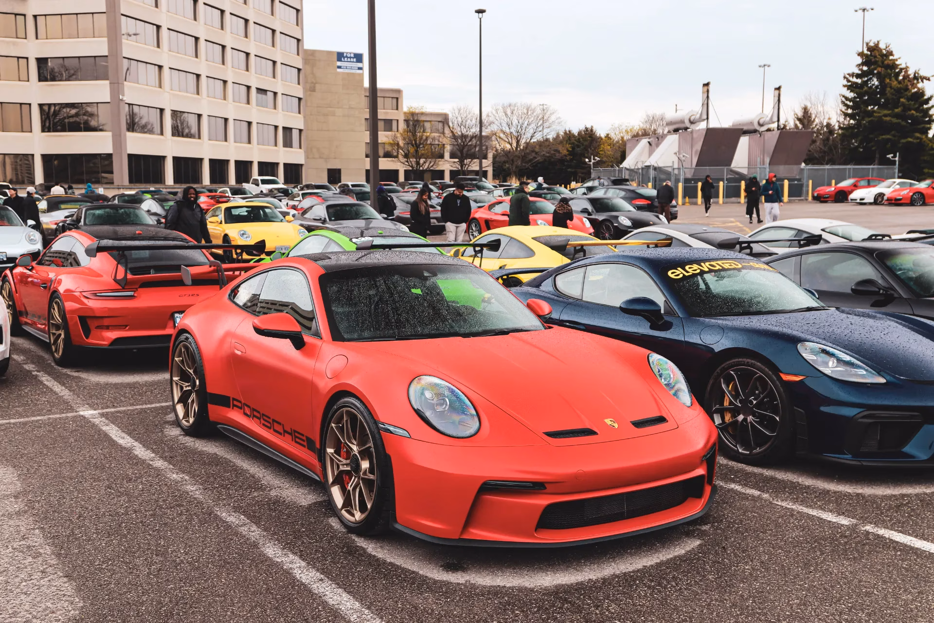 A red Porsche 911 with bronze wheels at a large parking lot meet, dozens of Porsches behind
