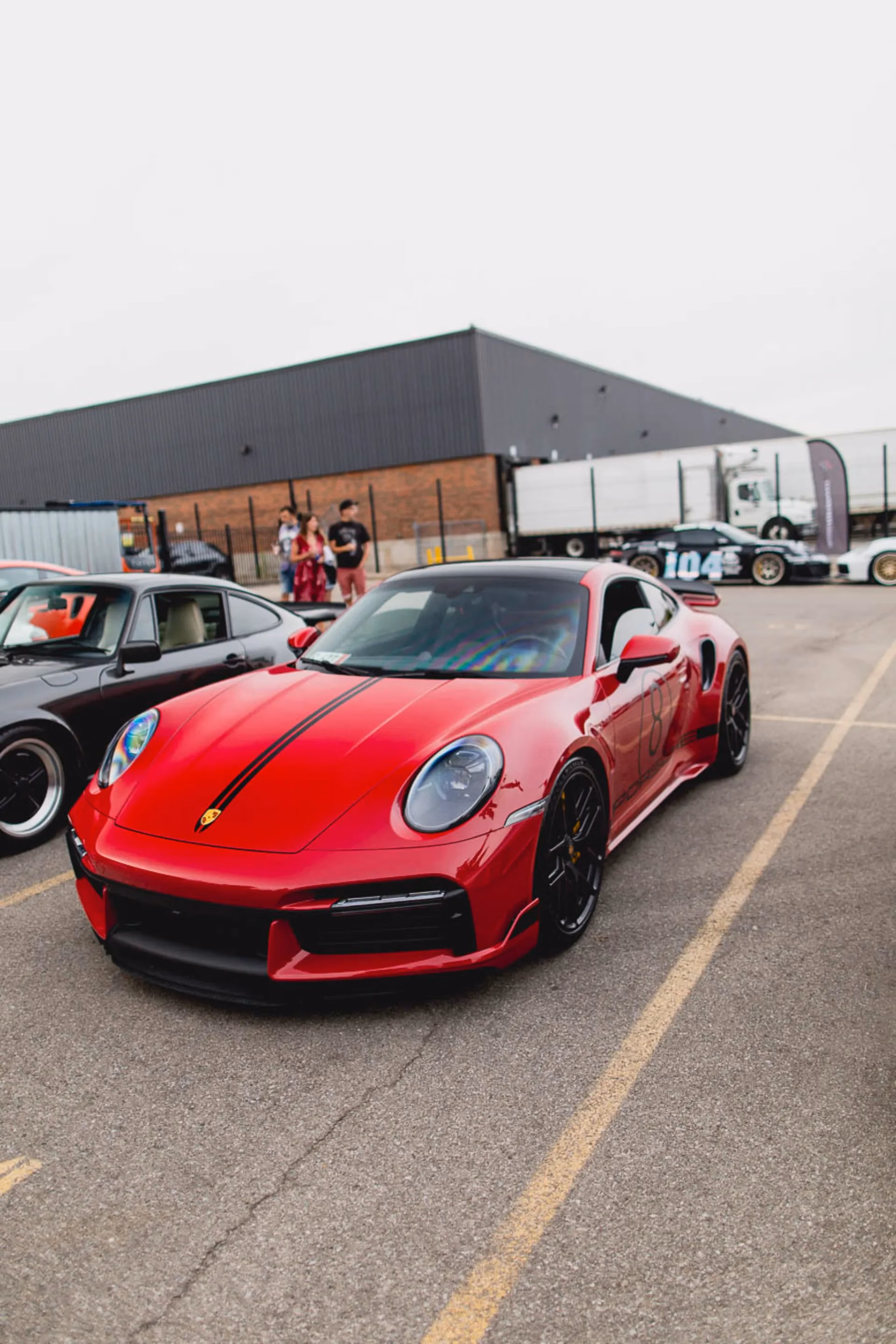 A red Porsche 911 parked at an outdoor meet, transport trailers behind it