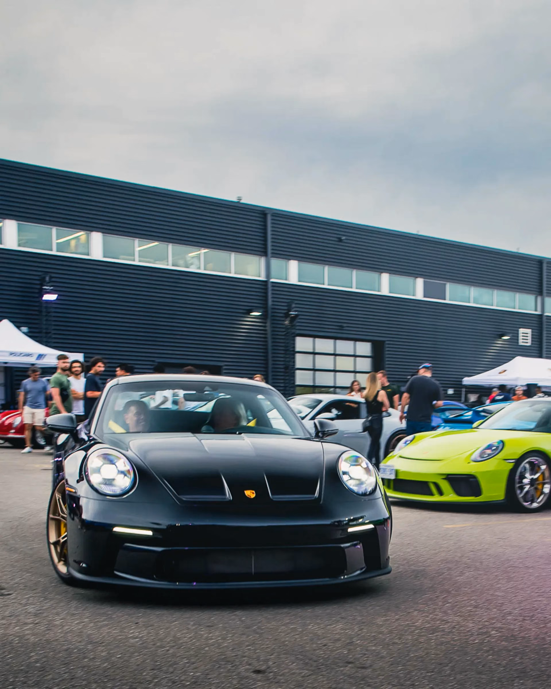 A black Porsche 911 with bronze wheels facing the camera at a meet, a lime green 911 alongside it