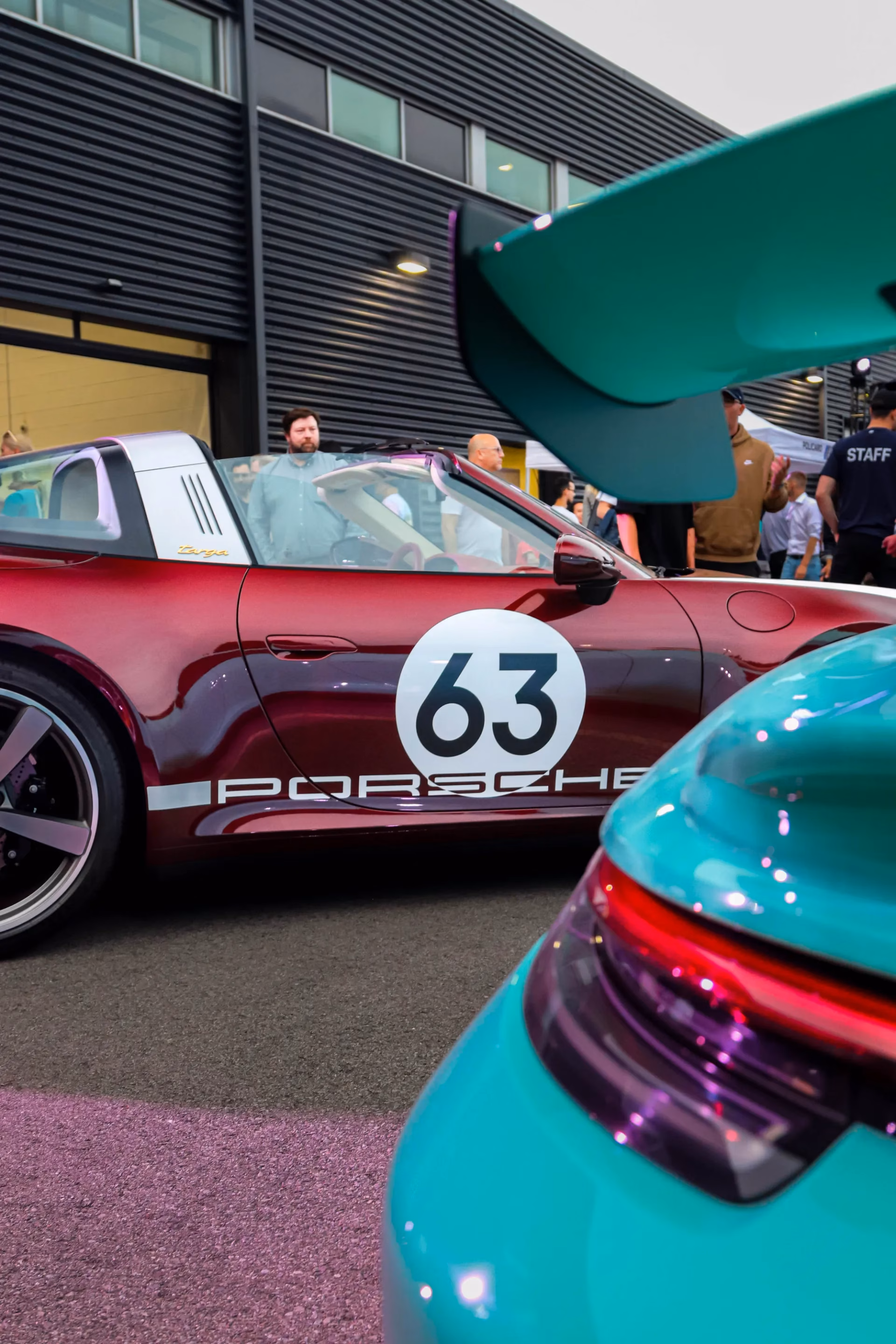A burgundy Porsche 911 Targa carrying a number 63 roundel at a meet, the rear wing of a green 911 in the foreground