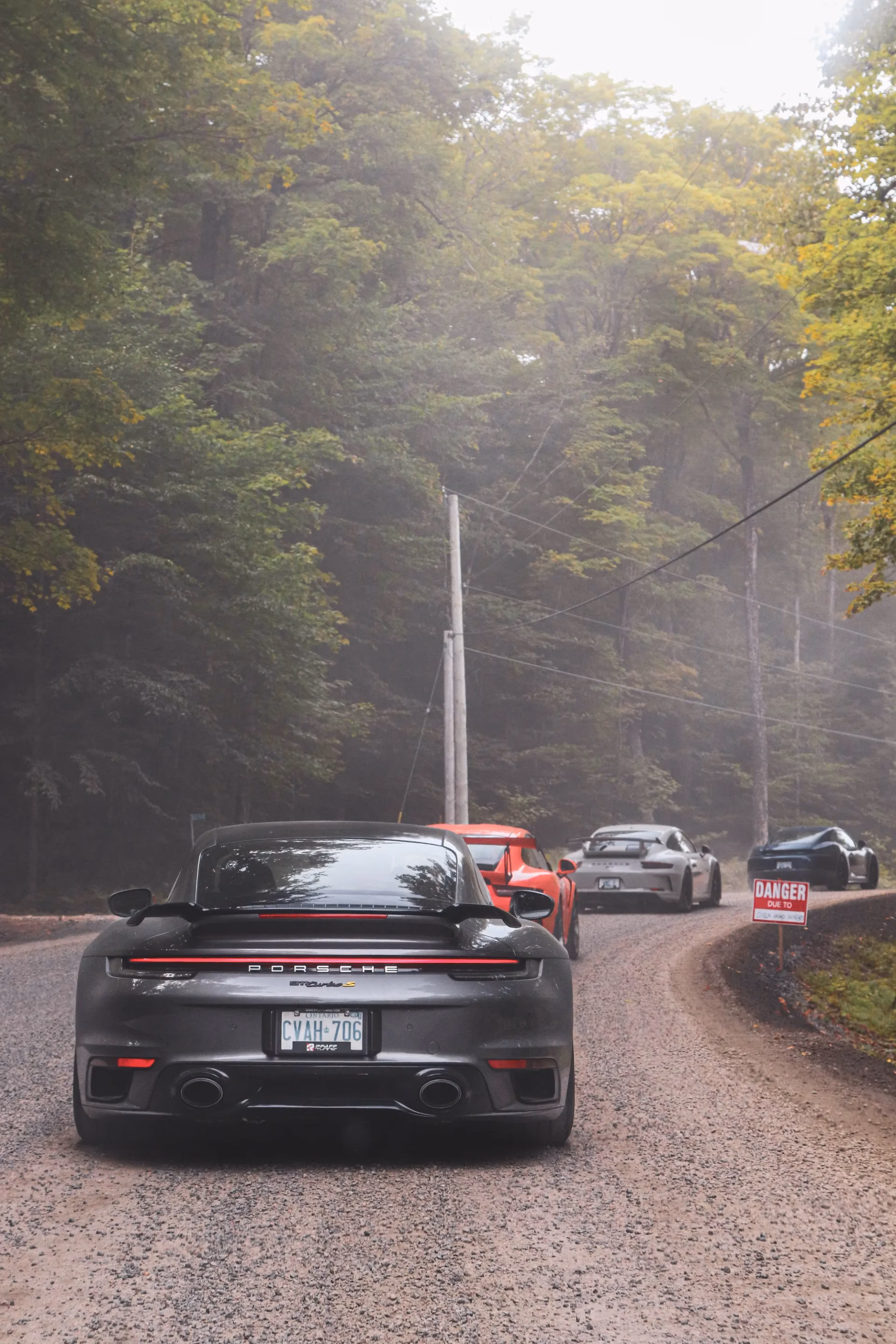 A dark grey Porsche 911 on a misty gravel forest road, more Porsches ahead
