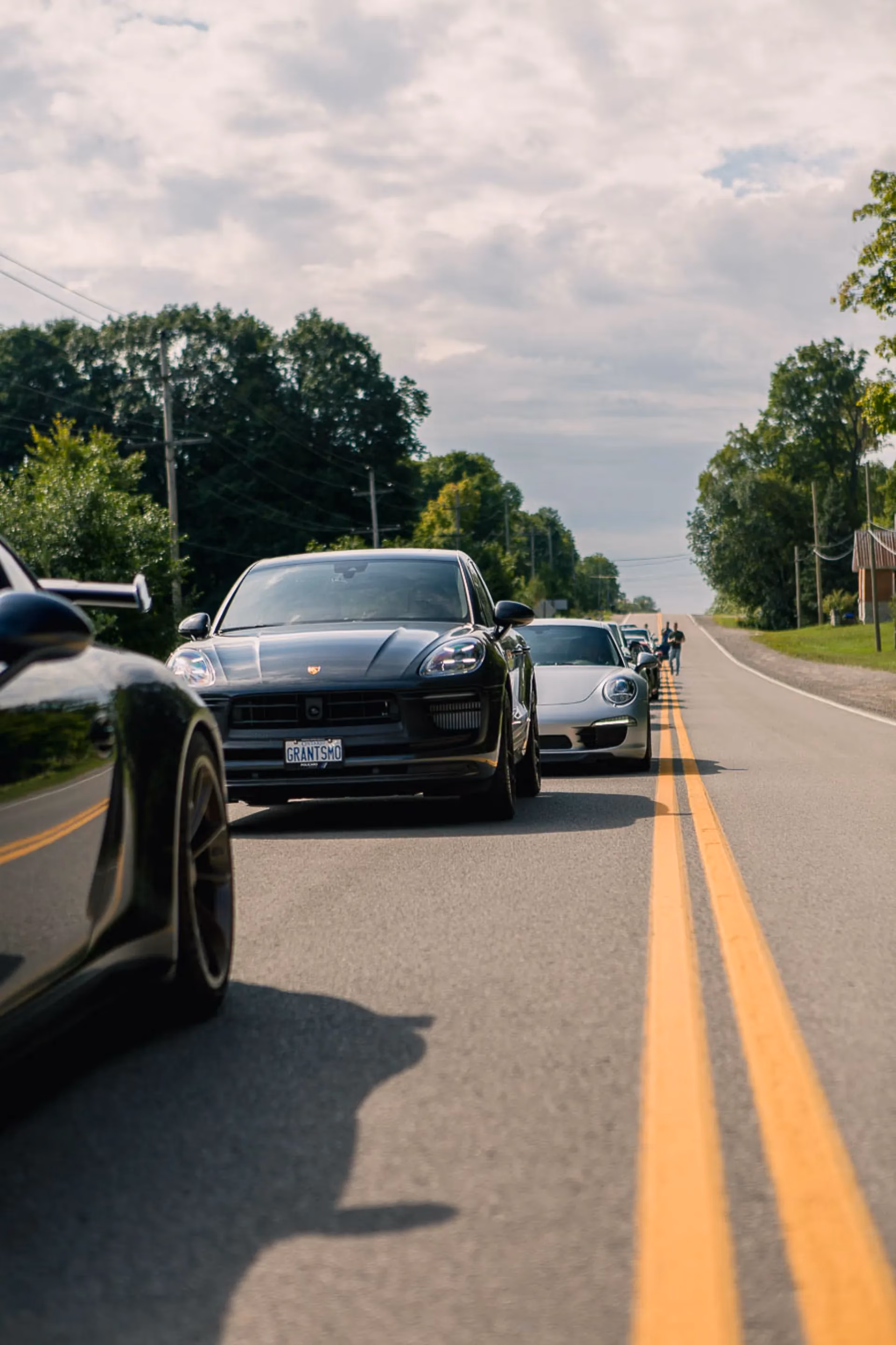 A black Porsche Macan leading a line of Porsches along a rural road