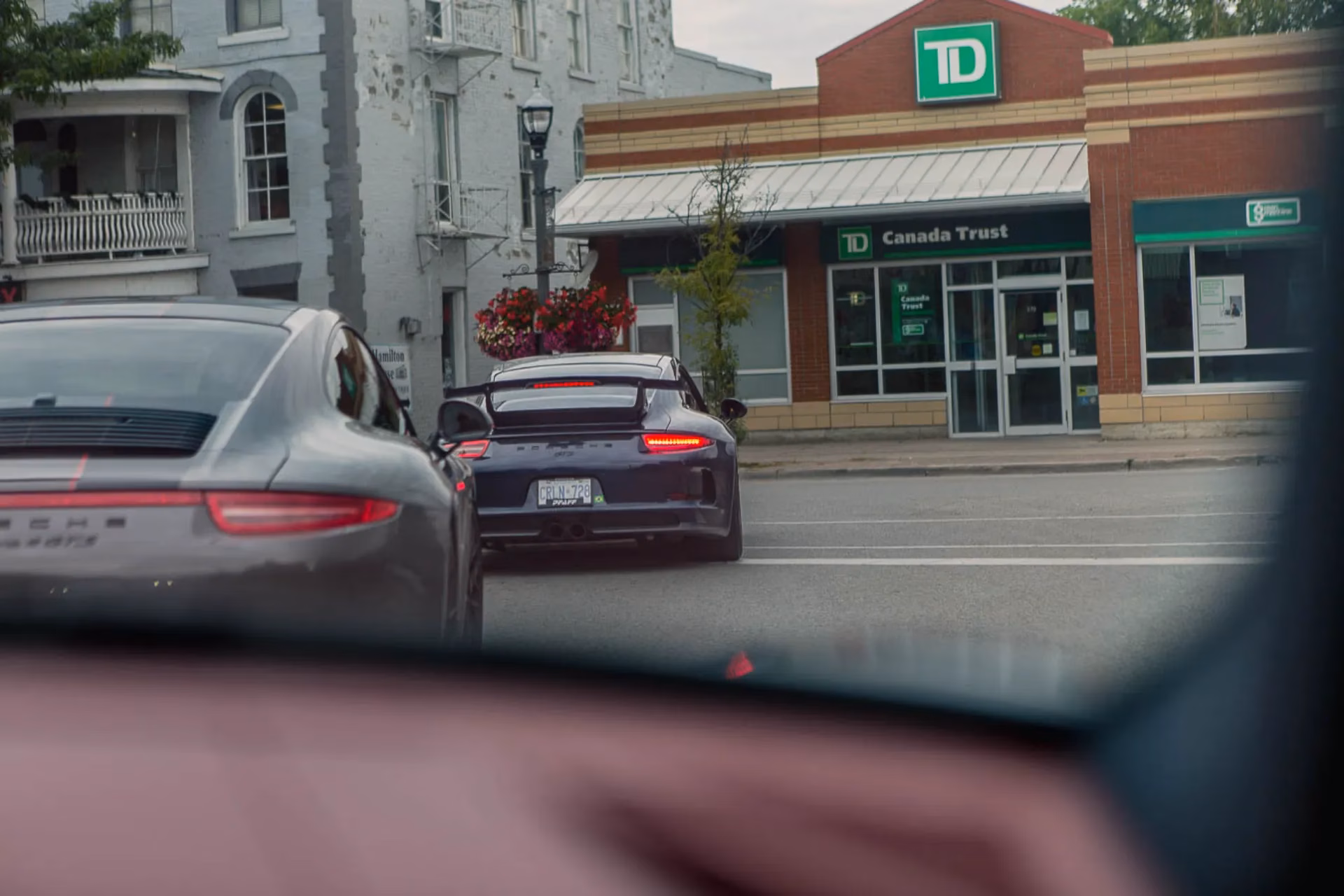 Two Porsche 911s driving through a small-town main street past a TD Canada Trust branch