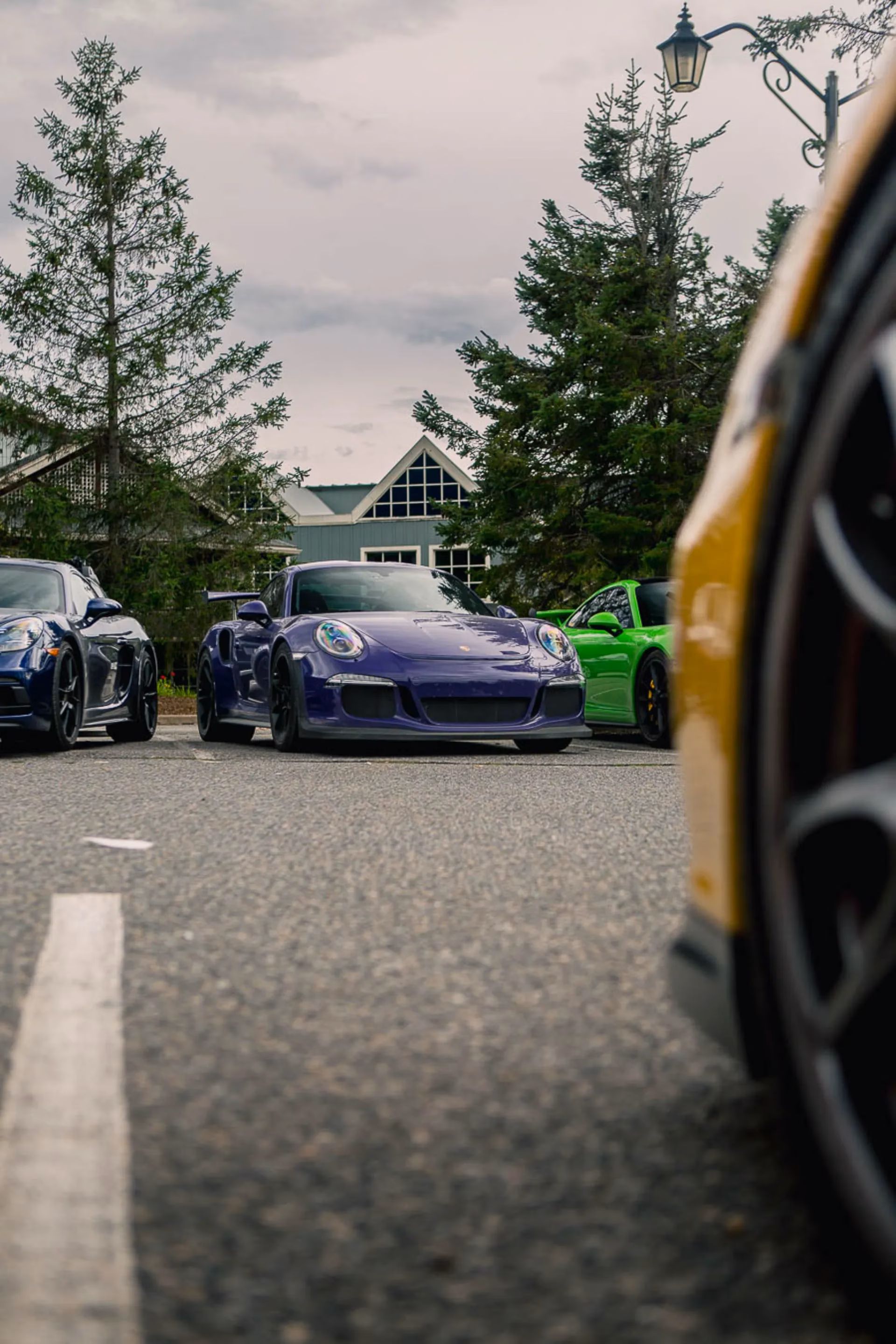 A purple Porsche 911 parked at a lodge, seen past the wheel of a yellow Porsche