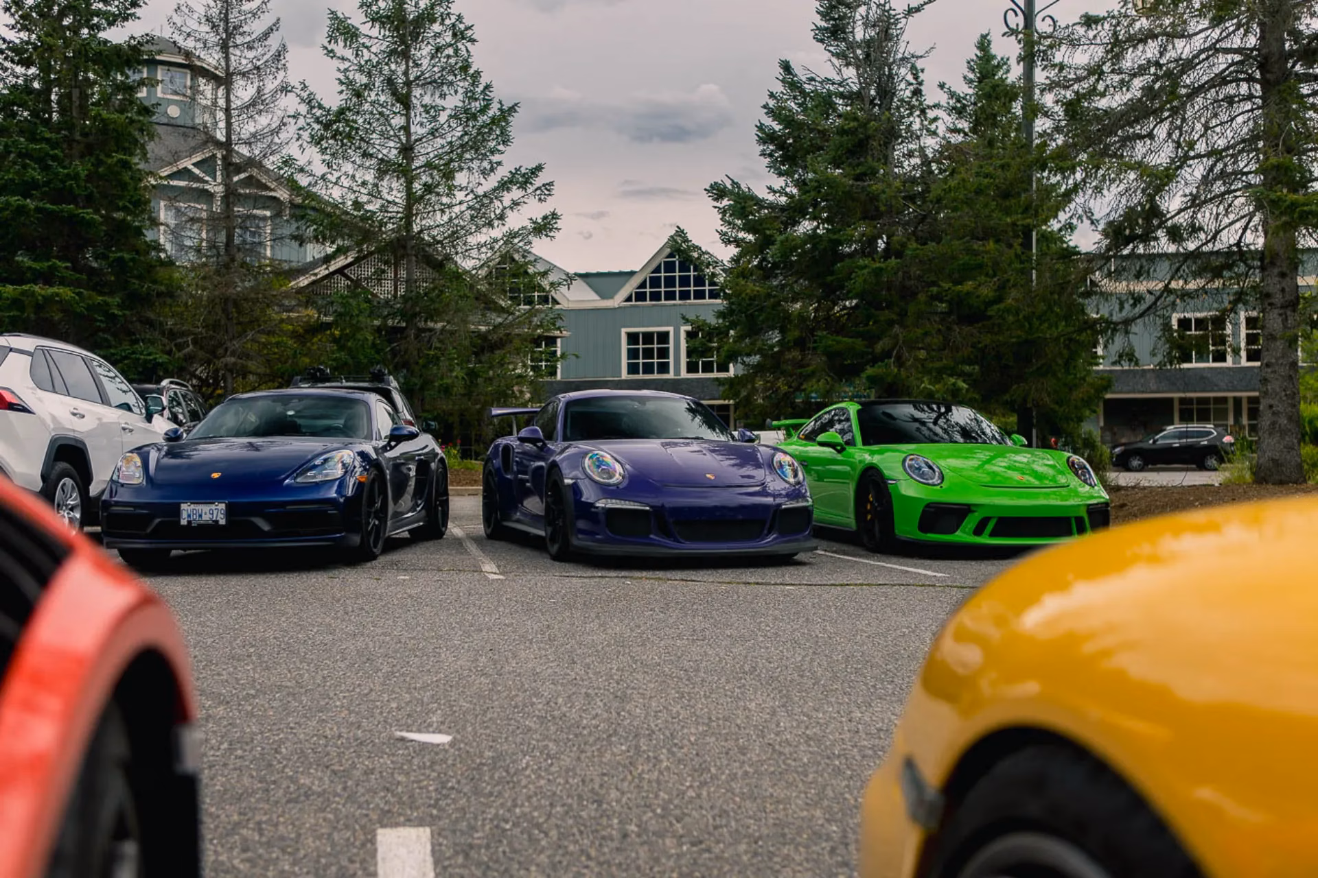 A blue Cayman, a purple 911 and a green 911 parked in a row outside a lodge
