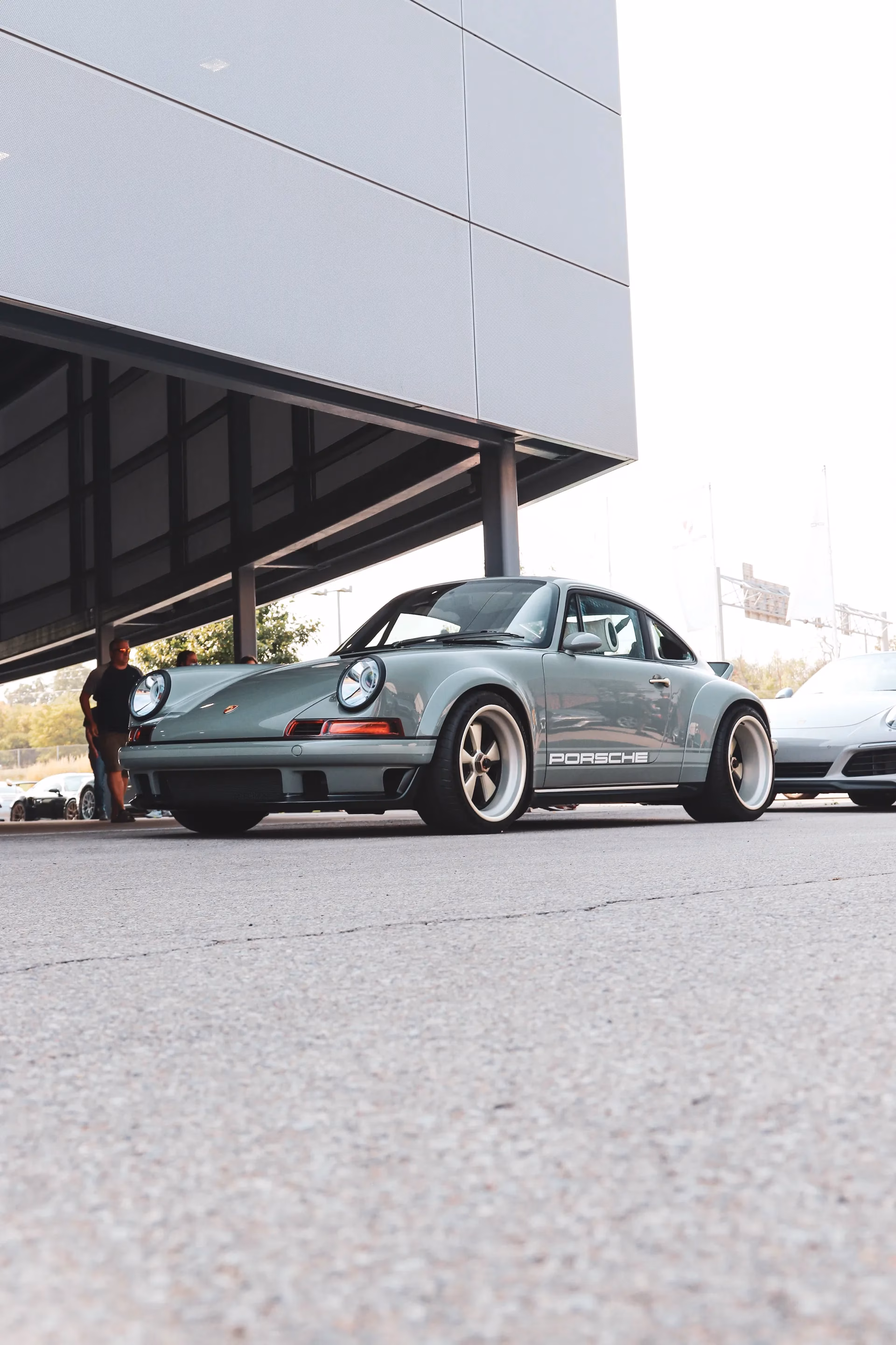 A grey-green restomod classic Porsche 911 with gold wheels parked beside a modern building