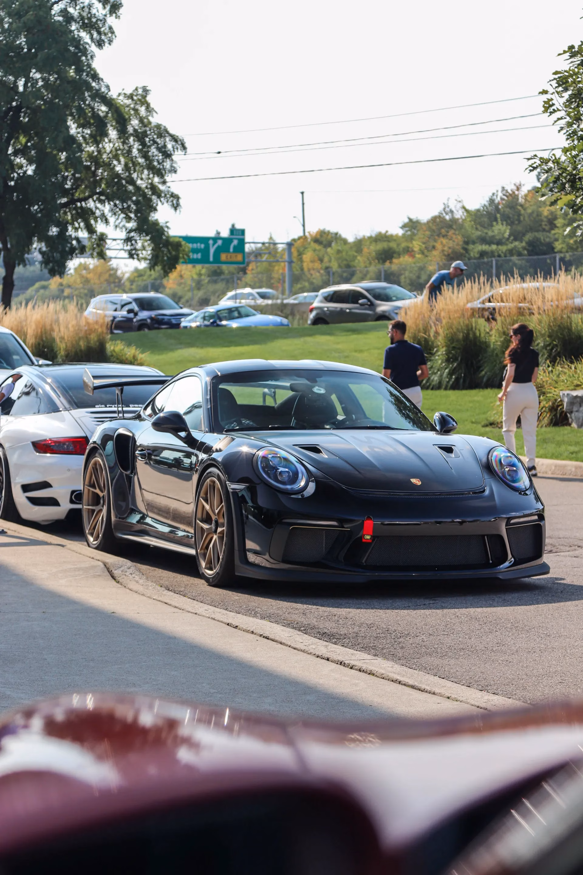 A black Porsche 911 with bronze wheels driving through a meet, a white 911 behind