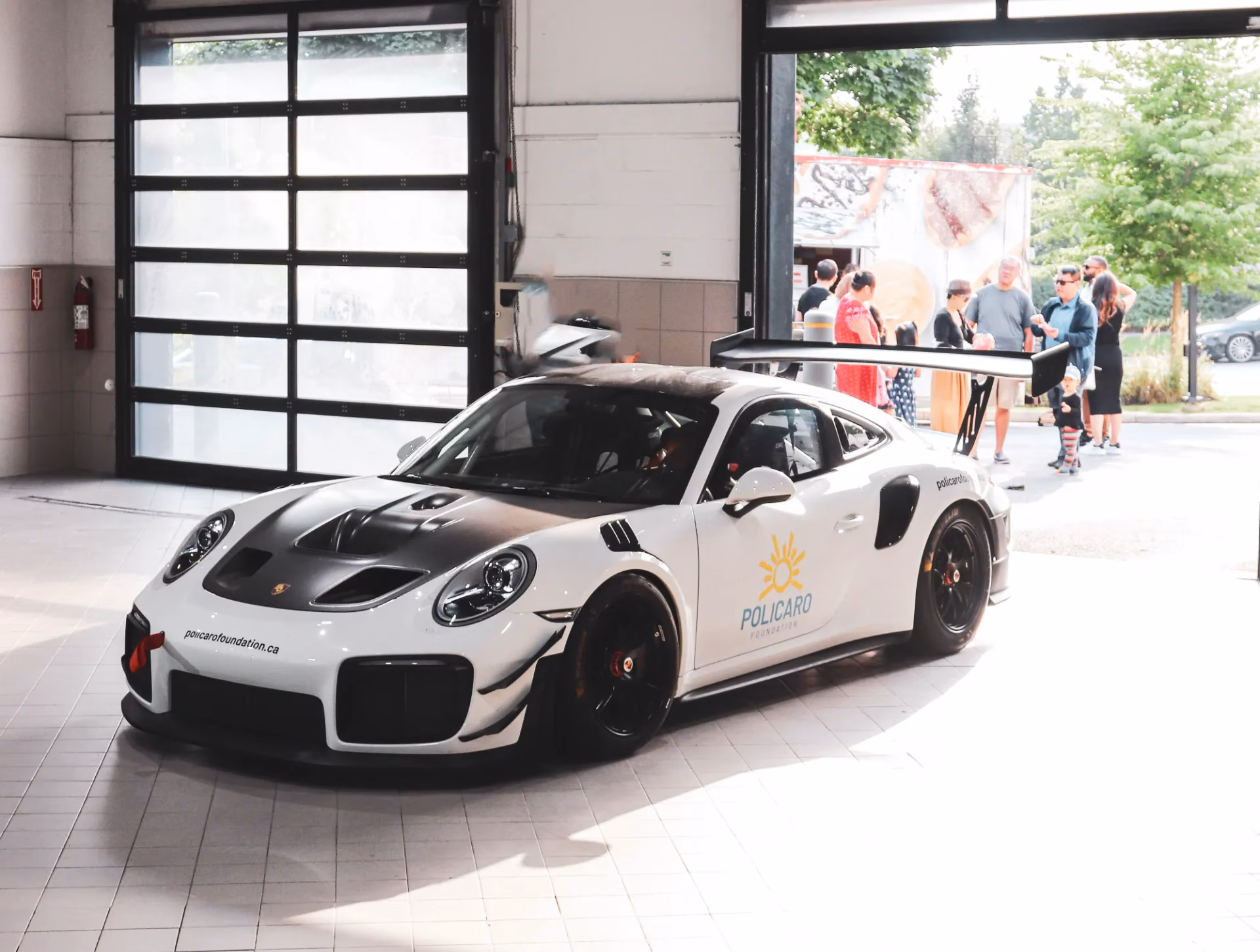 A white Porsche 911 race car in Policaro livery inside a service bay, members gathered behind