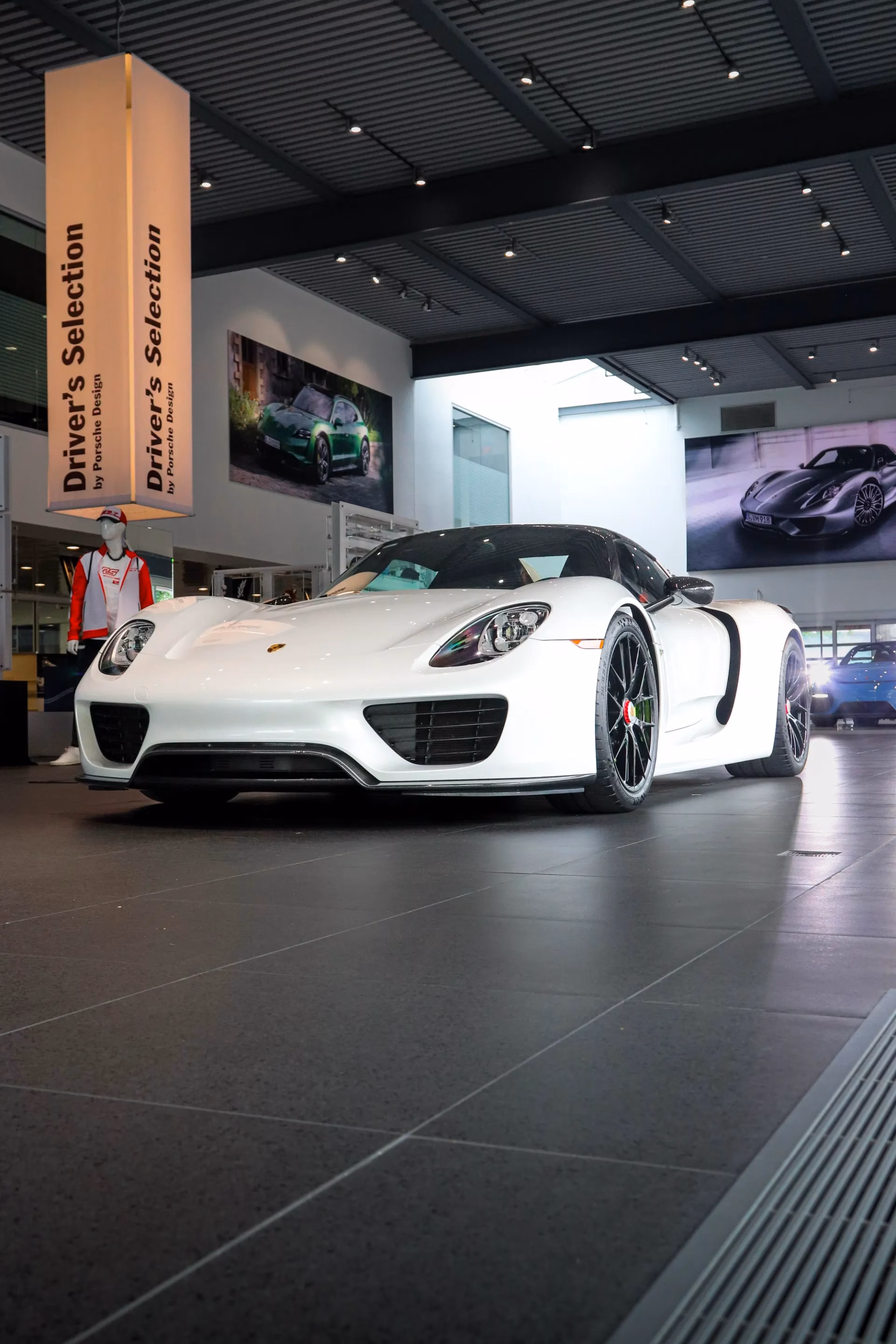A white Porsche 918 Spyder on display in a showroom beside Driver's Selection banners