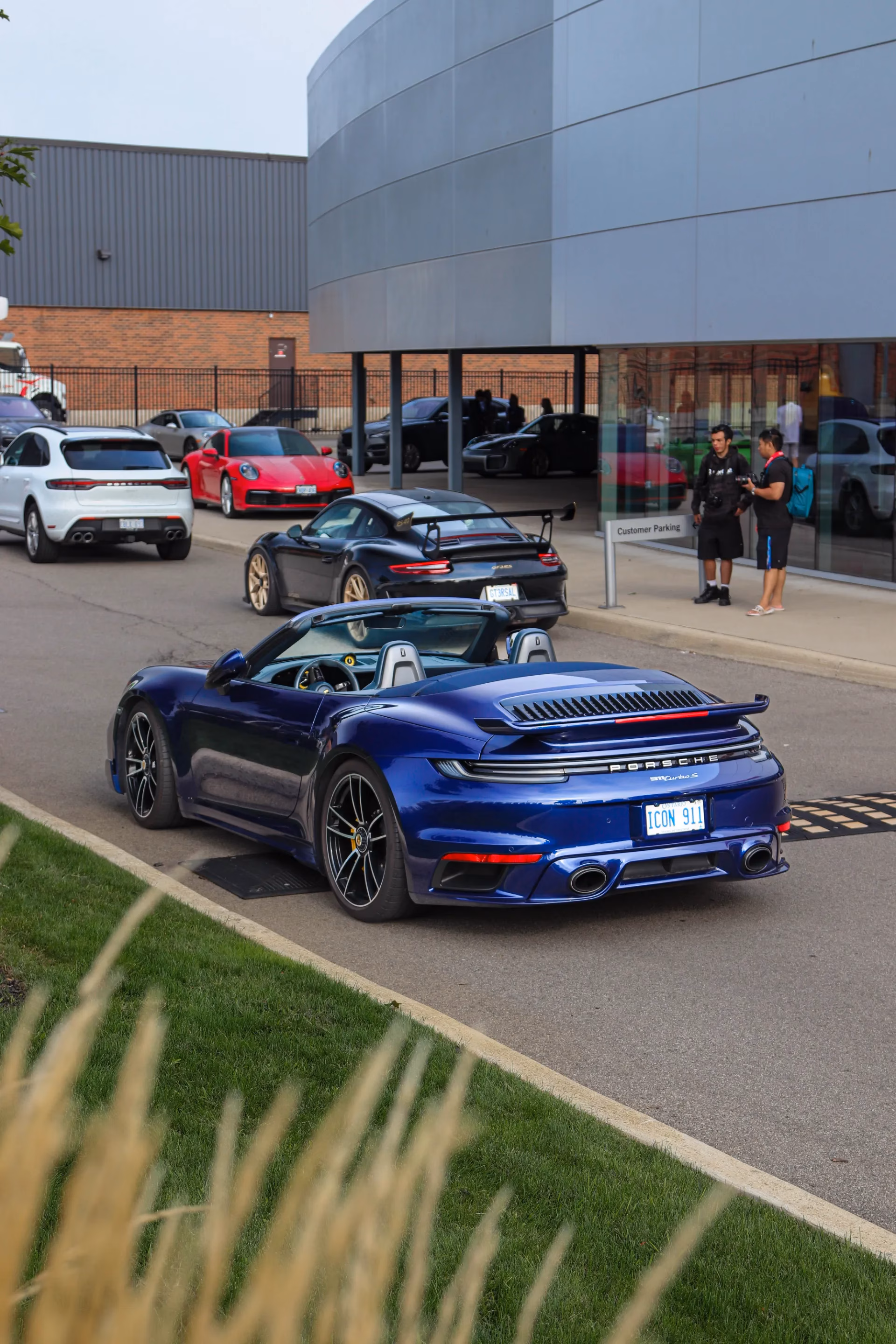 A blue Porsche 911 cabriolet parked outside a dealership, more Porsches behind