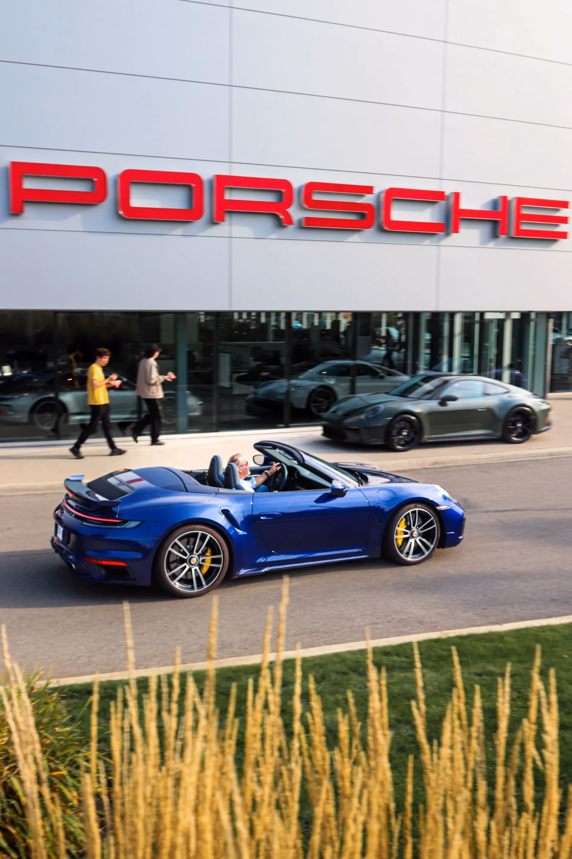 A blue Porsche 911 cabriolet driving past a Porsche dealership