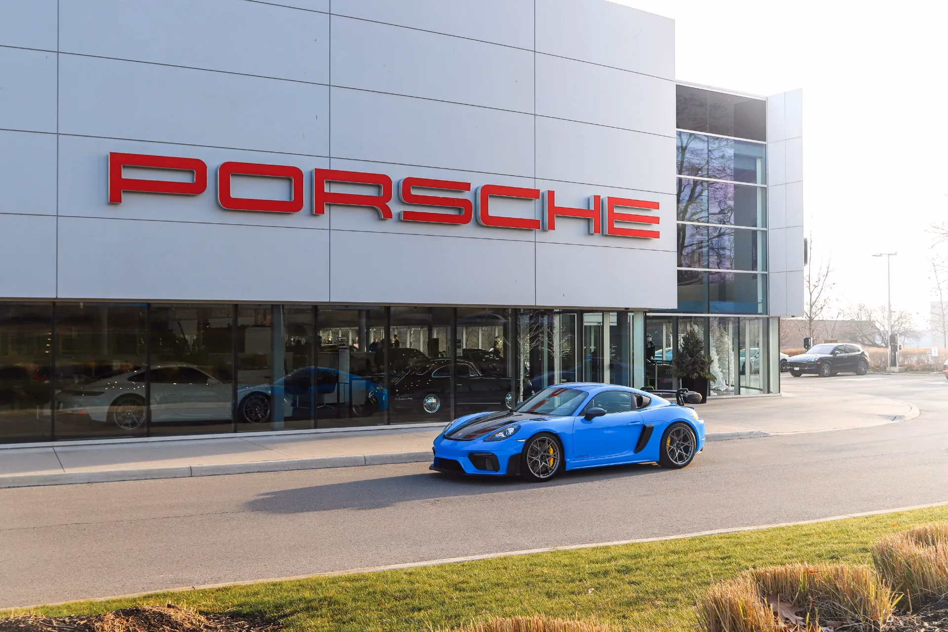 A blue Porsche Cayman GT4 RS parked outside a Porsche dealership beneath its sign