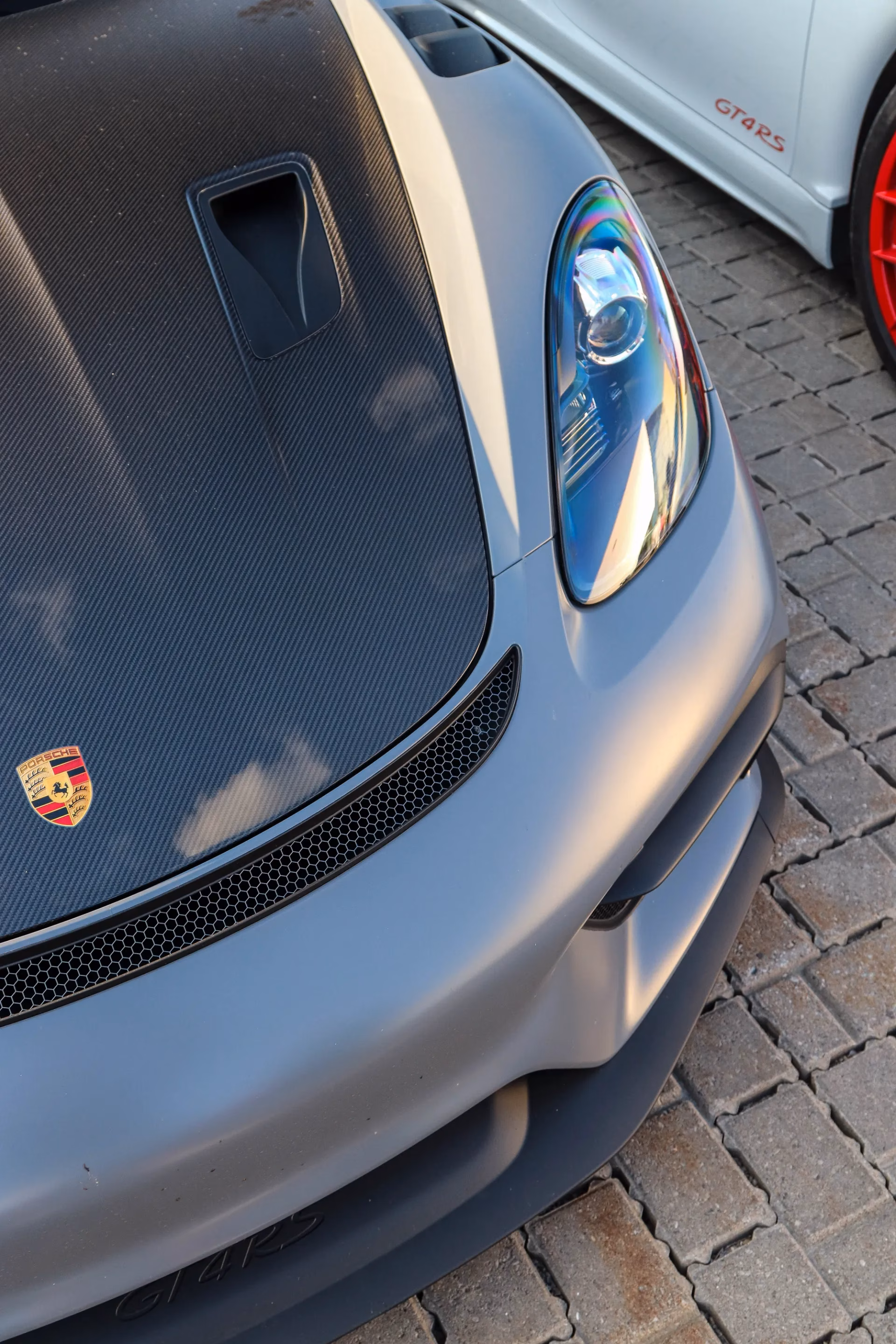 Close-up of a grey Porsche Cayman's carbon hood and crest, a white GT4 RS parked behind