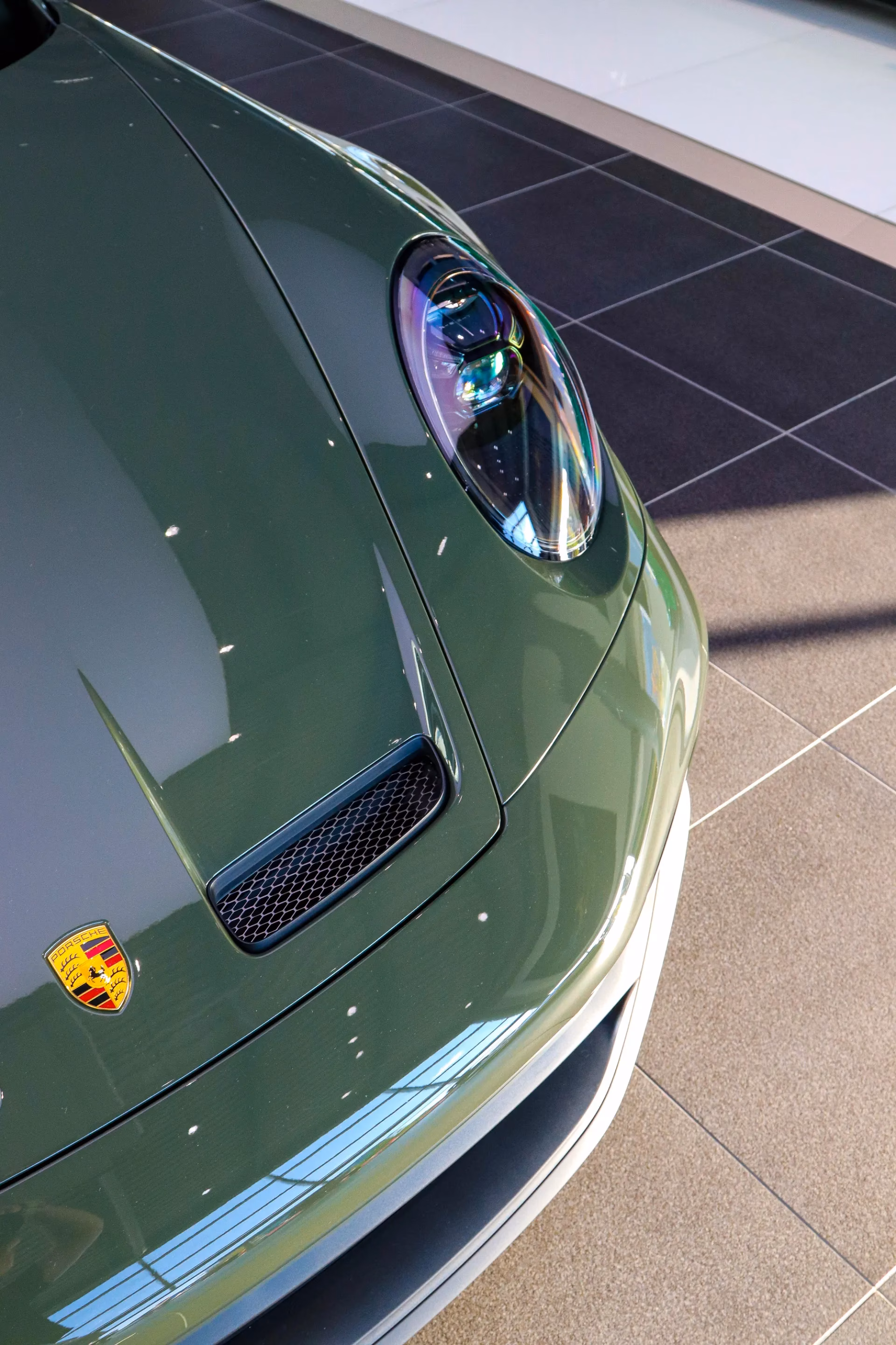 Close-up of a dark green Porsche 911's hood, crest and headlight in a showroom