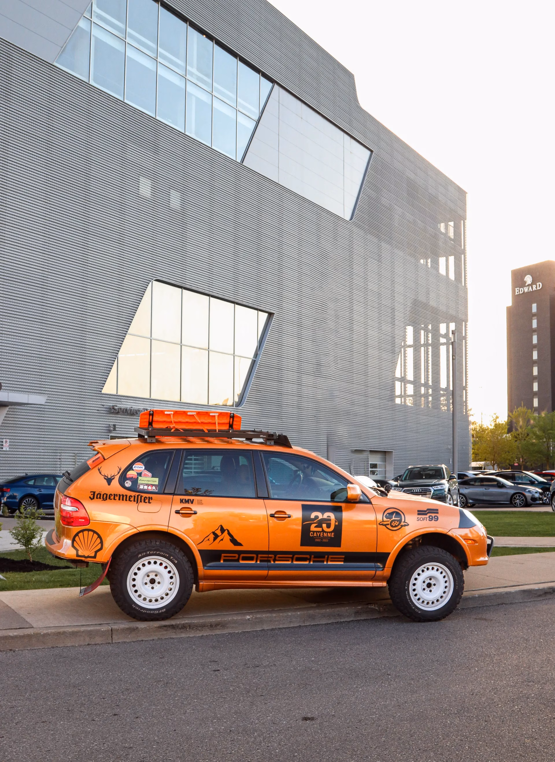 An orange Porsche Cayenne in rally livery with a roof rack and light bar parked outside a modern building