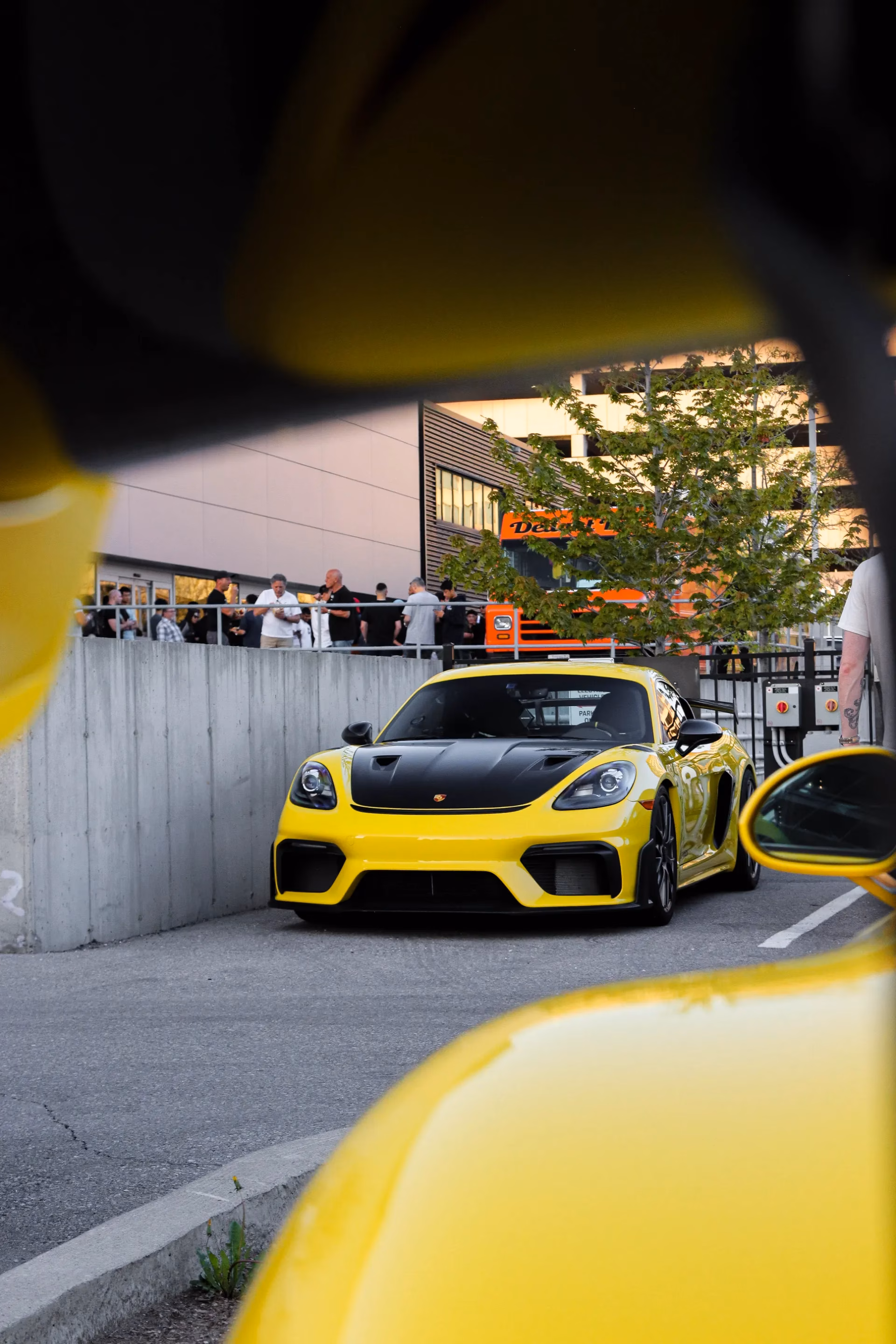A yellow Porsche Cayman framed between two other yellow cars at an evening club meet, members gathered on the terrace above