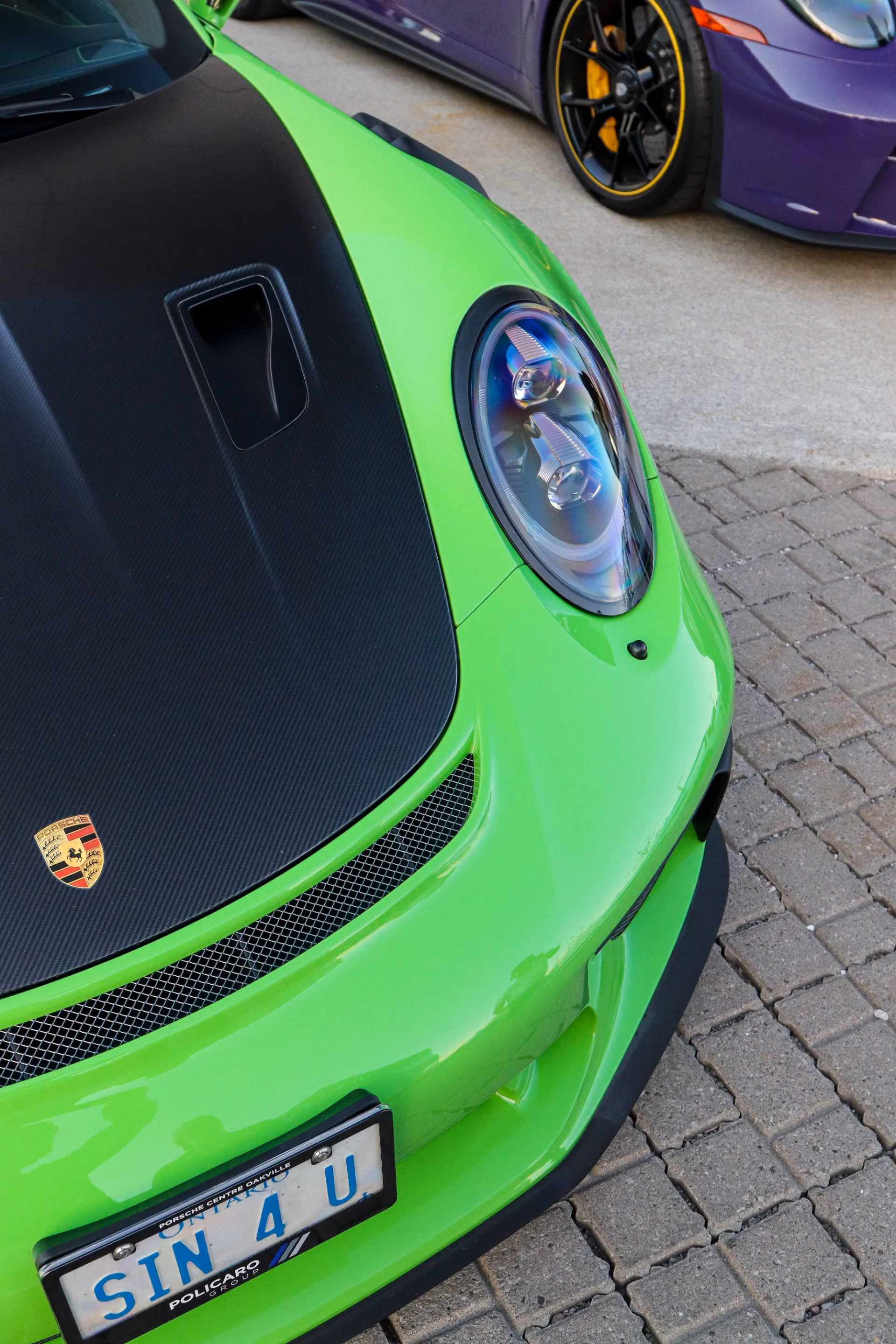 Close-up of a lime green Porsche 911's front end and Porsche crest, its licence plate reading SIN 4 U, with a purple Porsche parked behind