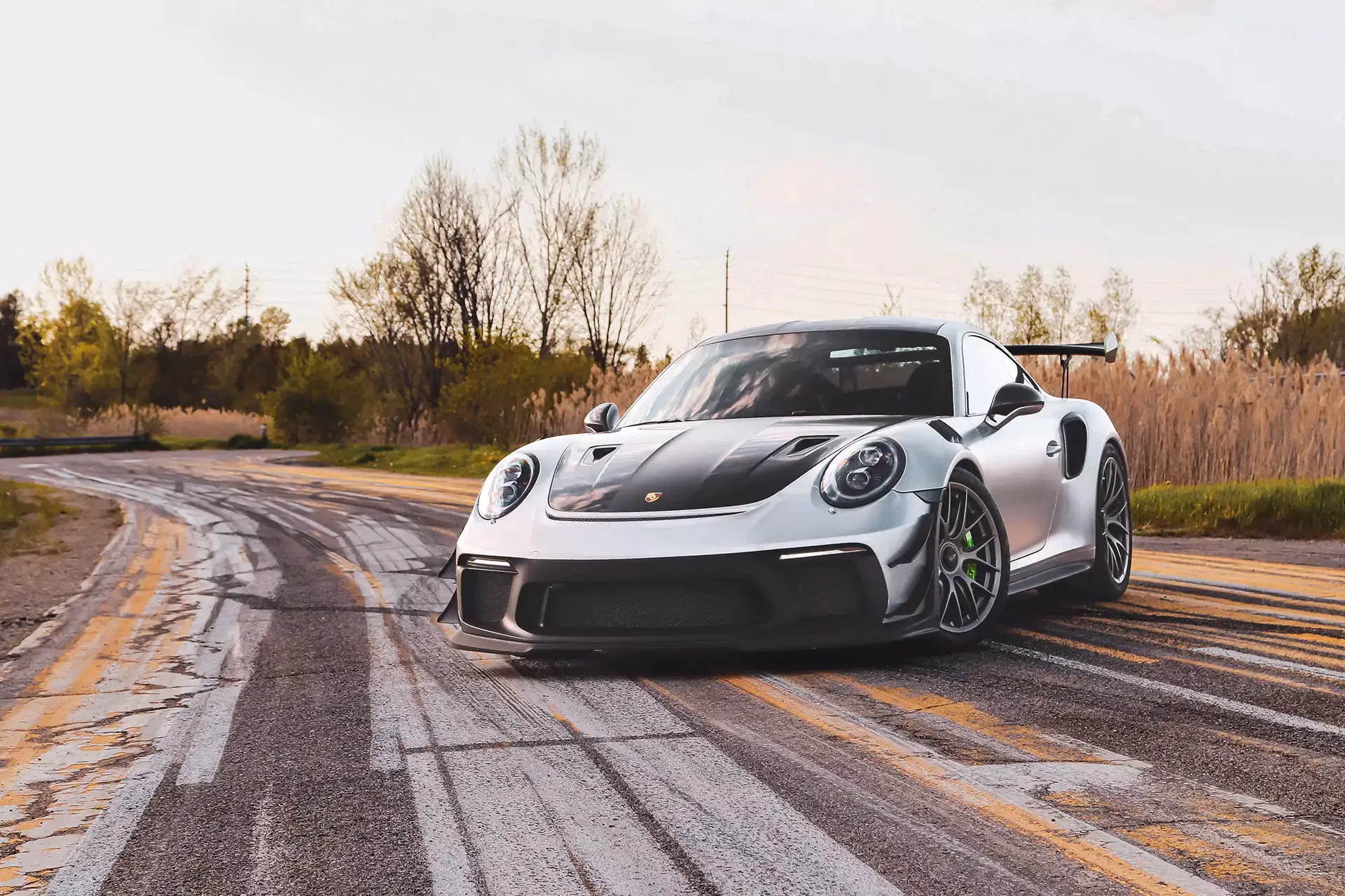 A silver Porsche 911 with a large rear wing on a country road at dusk