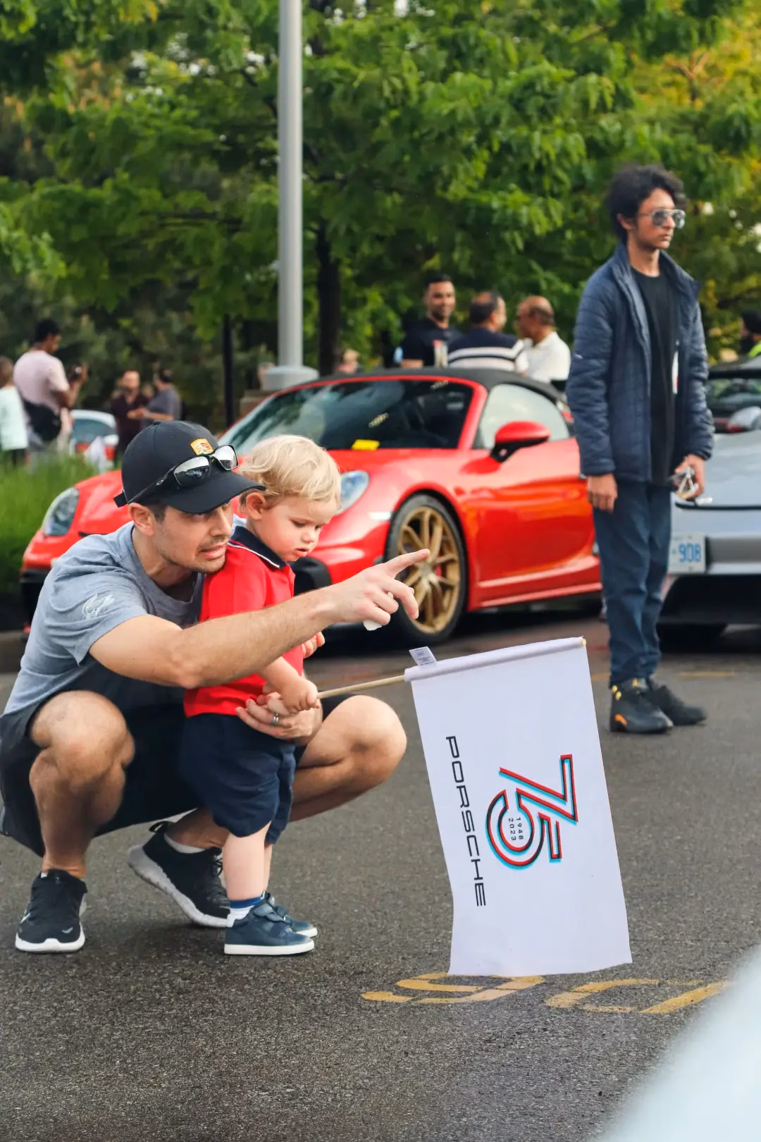 A member crouching with a small child beside a Porsche 75th anniversary flag, a red 911 parked behind