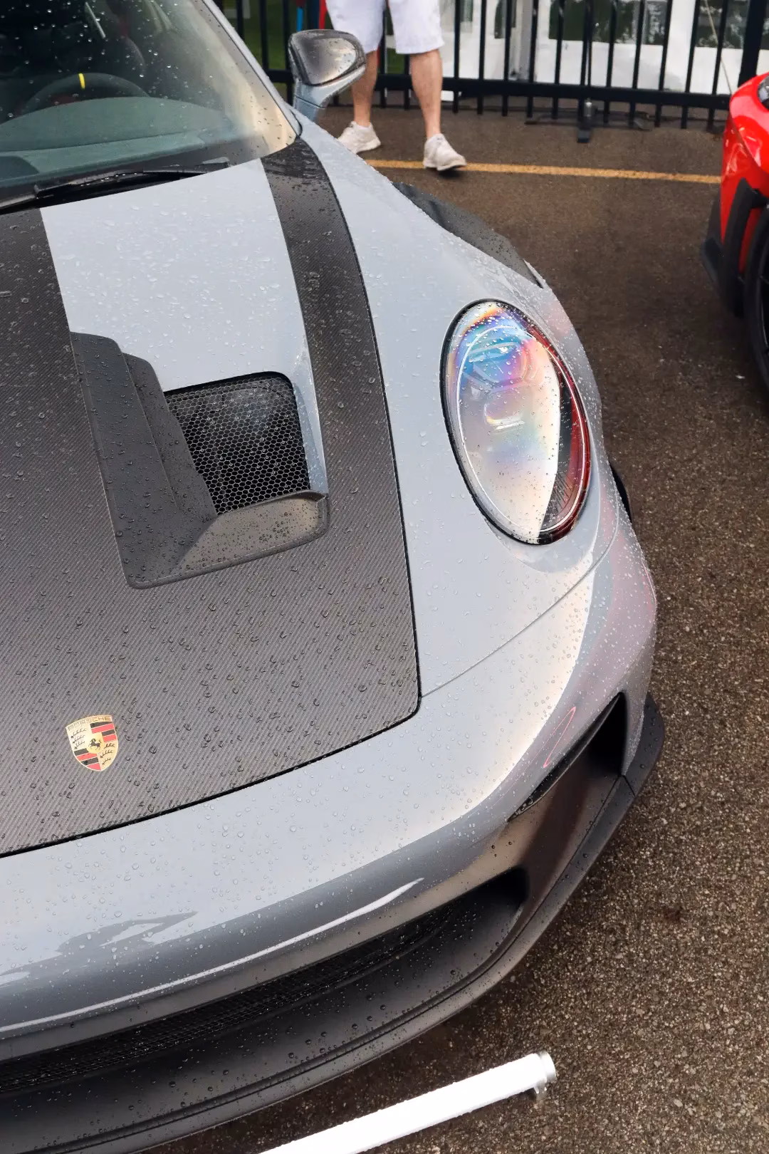 Close-up of a chalk grey Porsche 911's hood, headlight and crest beaded with rain