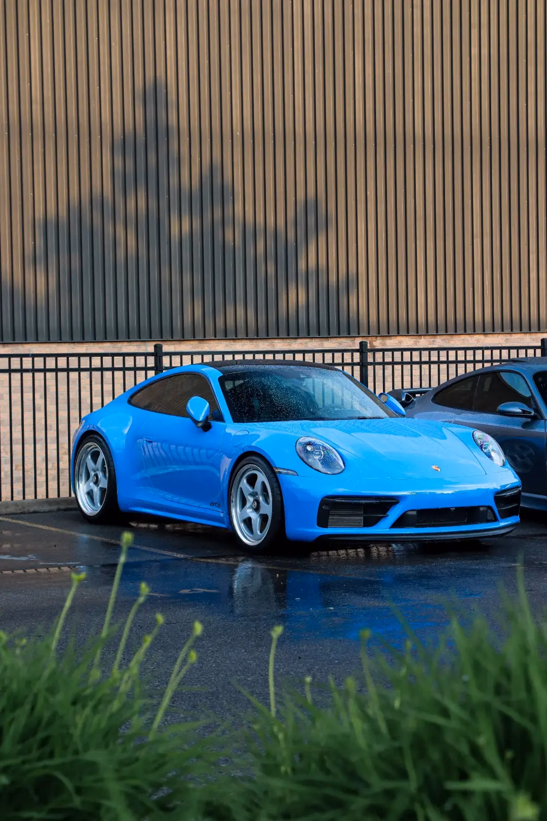 A bright blue Porsche 911 parked beside a fence at a rain-soaked meet