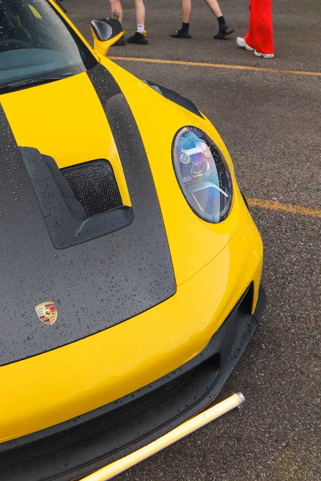 Close-up of a yellow Porsche 911's carbon hood and crest, members walking past behind