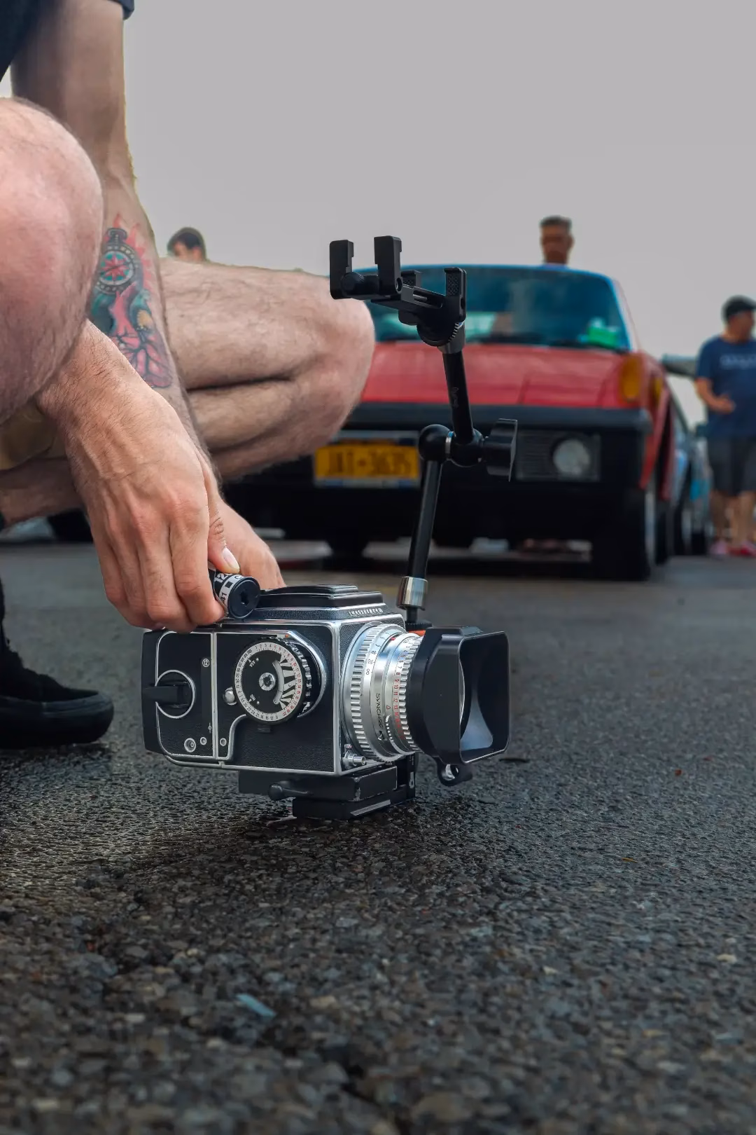 A photographer setting up a medium-format film camera low to the ground, a red classic car behind