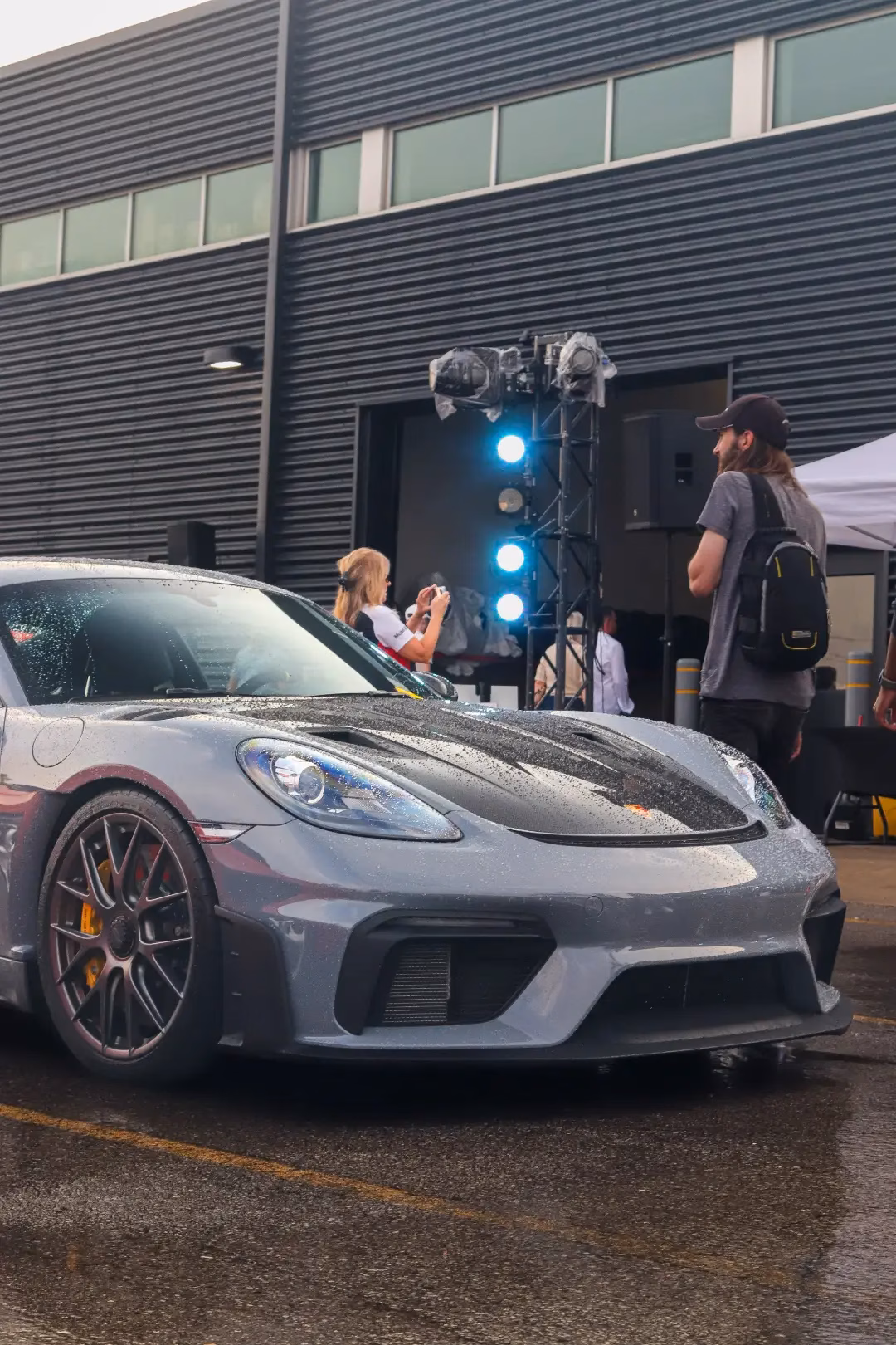 A grey Porsche Cayman at an evening meet, stage lighting and a photographer behind it