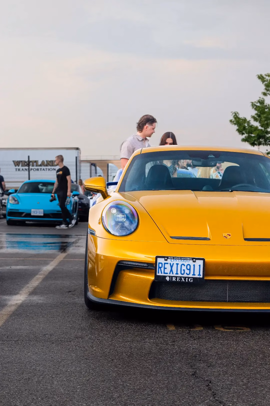 A yellow Porsche 911 with a REXIG911 licence plate at an evening meet, a blue Cayman parked behind