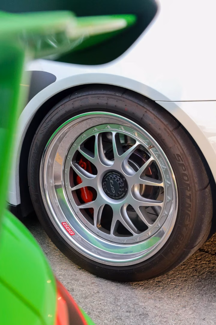 Close-up of a white Porsche's wheel and red brake caliper, a green car in the foreground