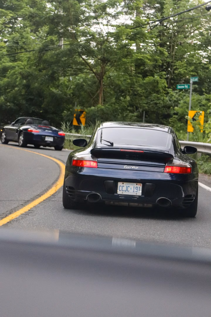 A dark blue Porsche 911 Turbo on a winding road, a black convertible ahead