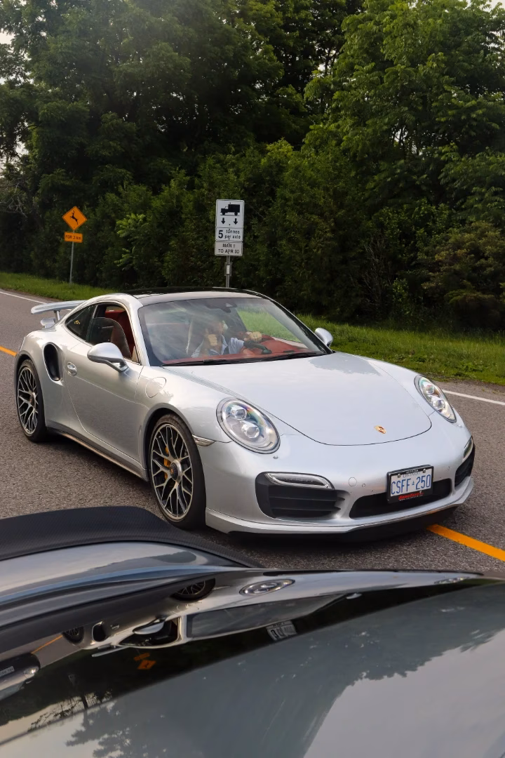 A silver Porsche 911 Turbo S on a rural road, driver and passenger visible inside