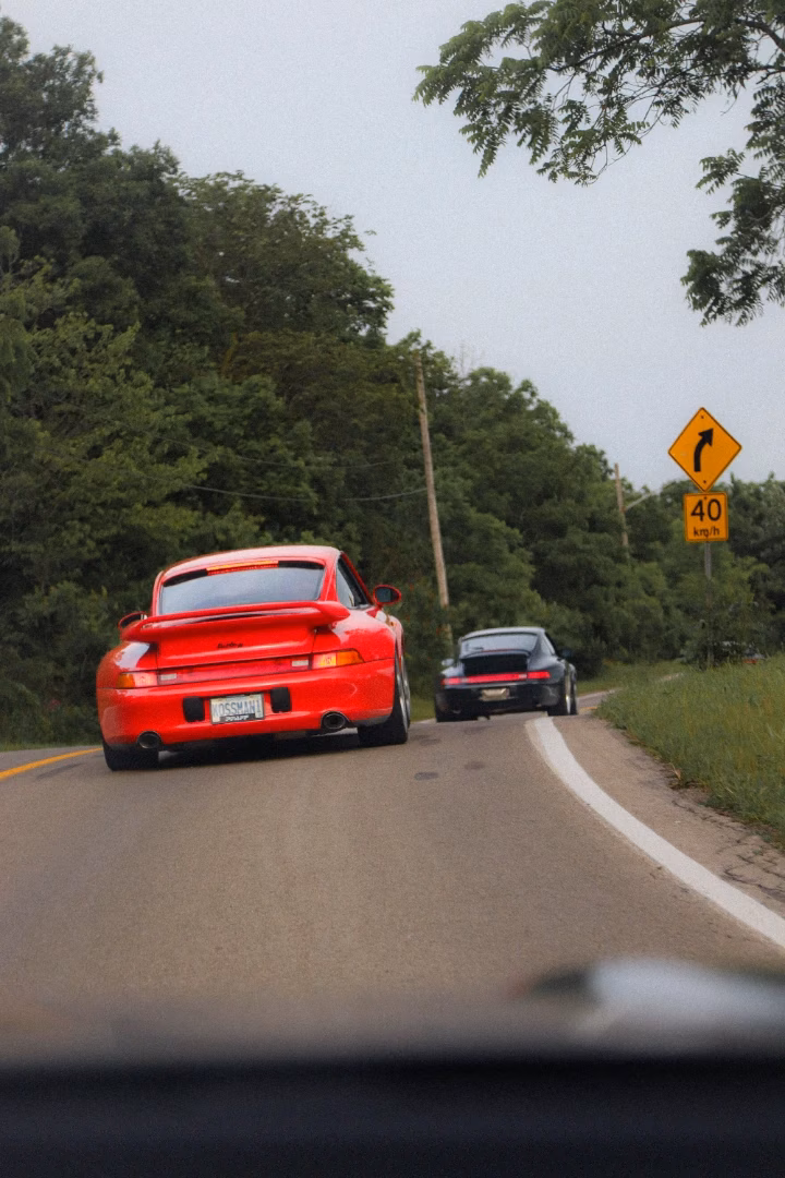 A red classic Porsche 911 following a dark Porsche along a country road