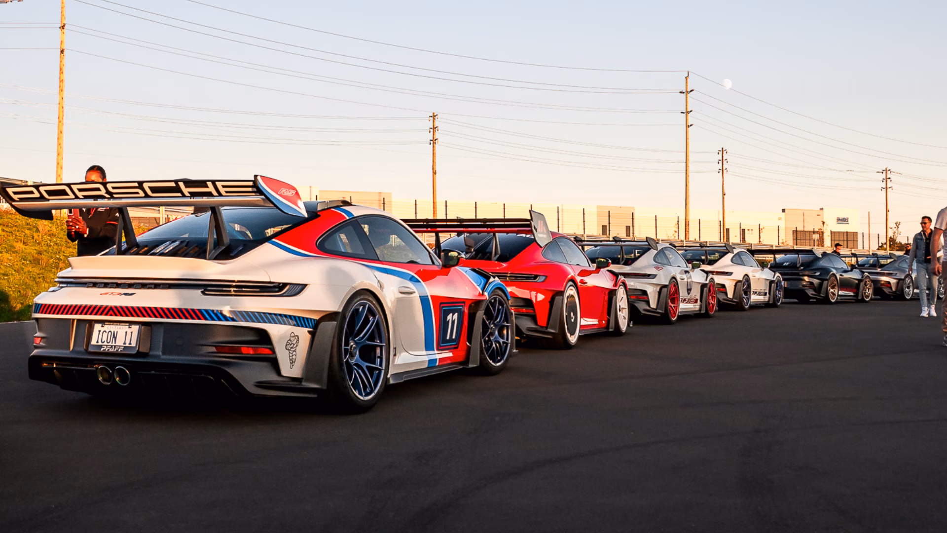 A white Porsche 911 in red and blue racing stripes at the head of a line of Porsches outside a dealership at sunset