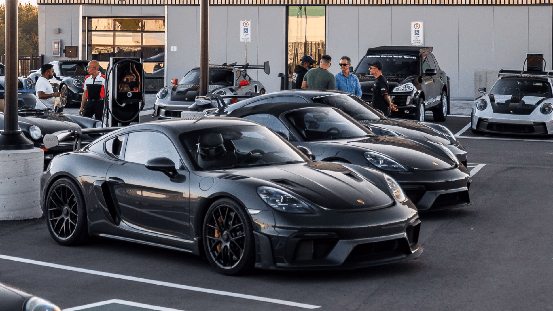 A dark grey Porsche Cayman parked at a morning cars and coffee meet outside a dealership