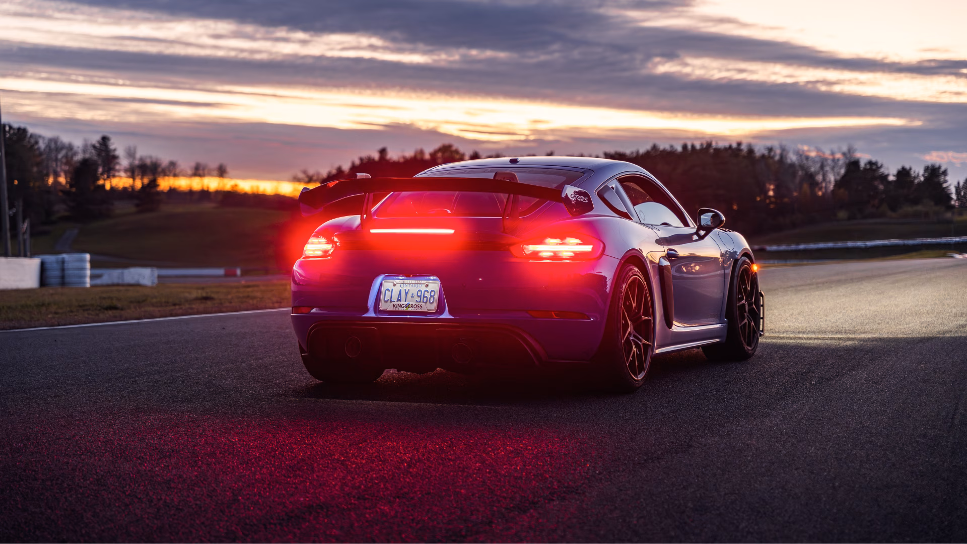 Rear view of a Porsche Cayman on a racetrack at sunset, tail lights glowing against an orange sky