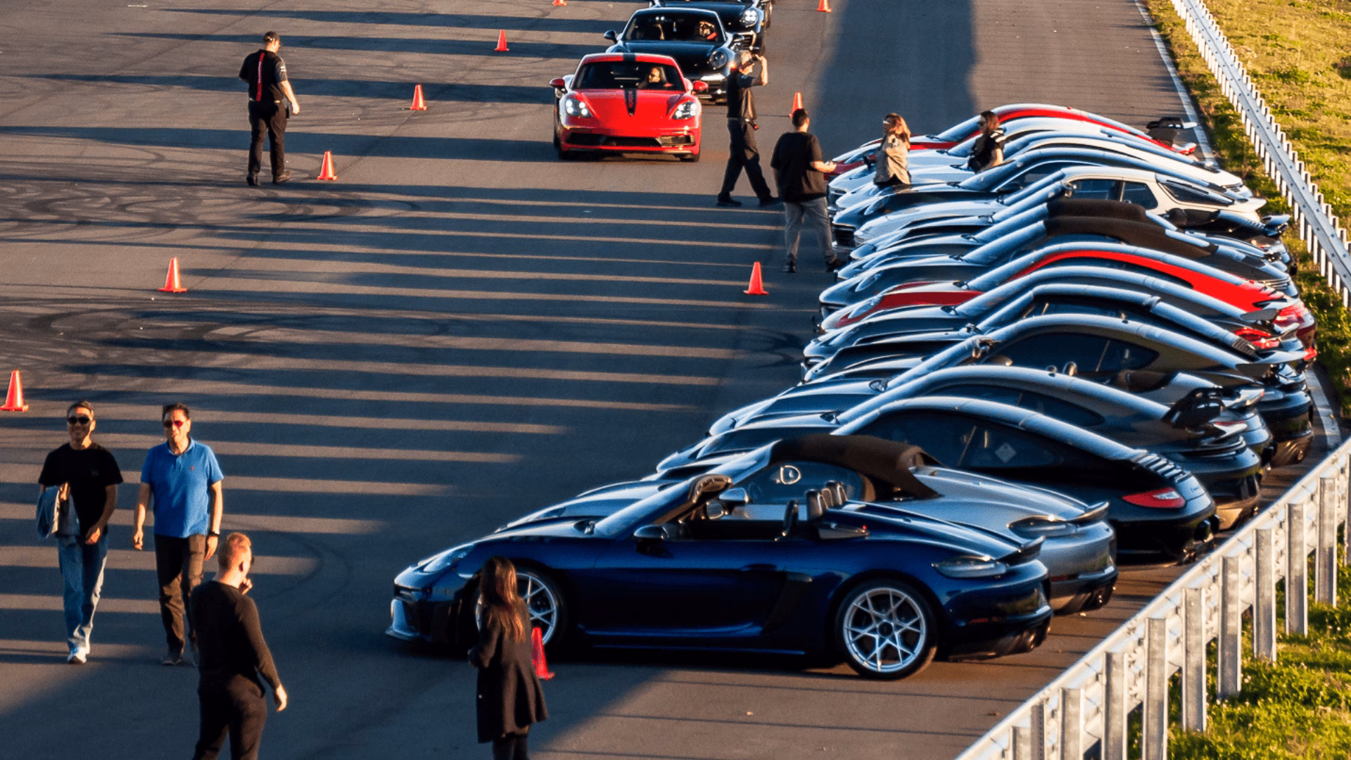 A long row of cars parked at an evening event, members walking between them in low sun