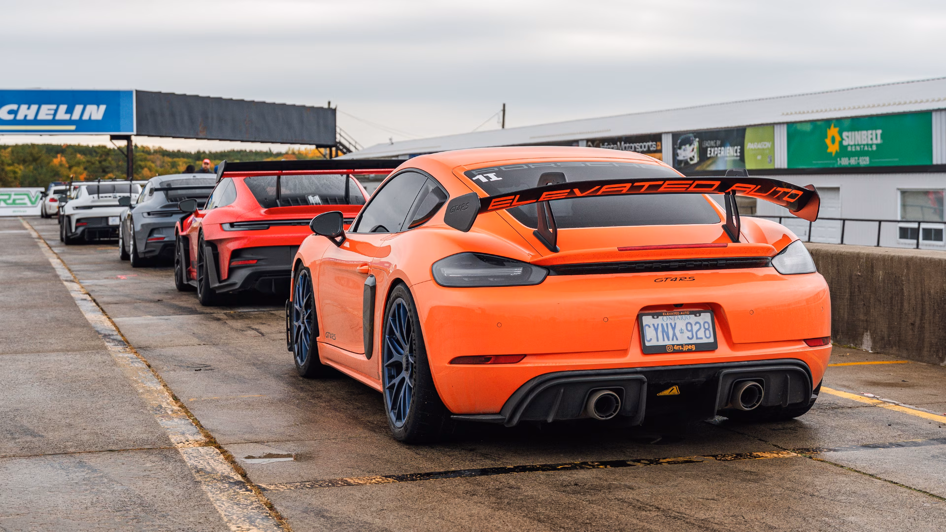 An orange Porsche Cayman with a large rear wing waiting in the pit lane, more cars lined up ahead beneath a Michelin banner